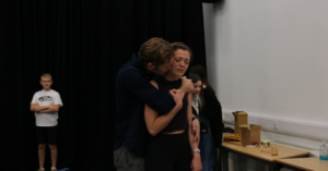A man stads hugging a woman from behind. The woman has a pained expression