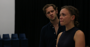 A woman looking teary faces forwards. A man standing at the side of her looks at her intently