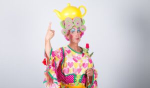 A man dressed as a panto Dame in bright clothing holding a rose and pointing to a yellow teapot on his head