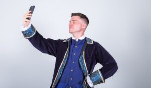 A man dressed as a prince holding out a phone taking a selfie