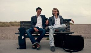An image of two men sat on a bench wearing waistcoats next to instruments in cases