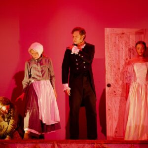 A woman crouches on the ground. A woman in a maid's costume stands reaching down to her next to a man in a suit who clutches his shoulder looking pained. A woman stands next to him in a white wedding dress