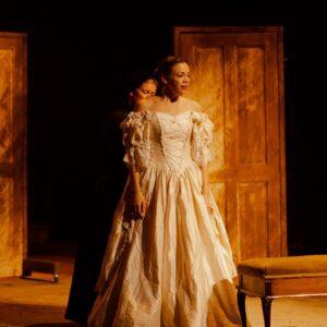 A woman stands in a white wedding dress, a woman in a black Victorian maid's dress stands behind, helping to do up her dress