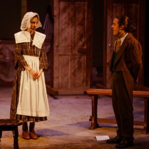 A young girl in Victorian dress and a bonnet stands with her hands in front of her looking anxiously at a man who wears a suit, with his hands behind his back
