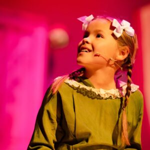 A young girl wearing a green dress and white bows in her hair looks up, smiling