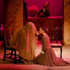 A woman in a brown dress kneeling before a person with a scarf draped over their head