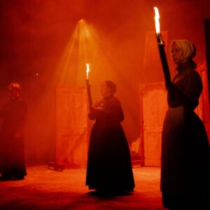 Two women in Victorian dress stand holding flaming torches in a red atmospheric light