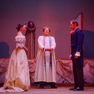 A man dressed as a priest stands talking in front of a woman in a white wedding dress and a man in a blue suit jacket and pink cravat. A woman in a long dress and nightgown stands behind them watching