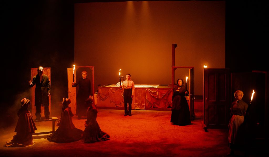 A group of people in Victorian dress stand onstage holding flaming torches in red atmospheric lighting