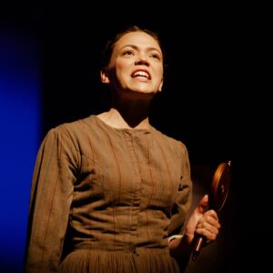 A close up of a woman in a brown striped dress talking passionately holding a mirror