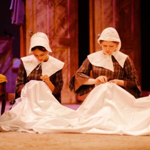 Two girls wearing brown checked dresses and white bonnets kneel whilst sewing