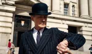 Bill Nighy stands in the street wearing a black suit and bowler hat. He is looking at his watch.