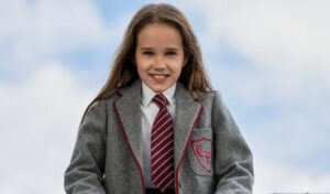 A young school girl wearing a grey school uniform smiles at the camera