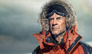 An image of Sir Ranulph Fiennes standing in an orange jacket with a fur lined hood looking out into the distance