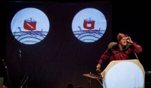 A man dressed as a pirate pretending to look through a telescope. Behind him two shadow puppets of boats are lit up against a cloth.