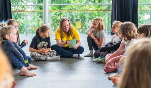 A group of children sat in a semi circle listening to a workshop leader who is reading out from a sheet of paper