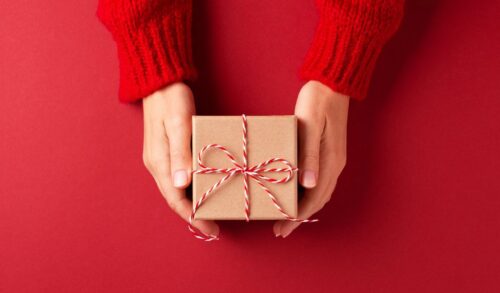Two hands holding a small square gift wrapped in brown paper with a red and white string bow against a dark red background