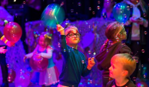 A young boy with his face painted is waving a balloon as bubbles fall down in front of him