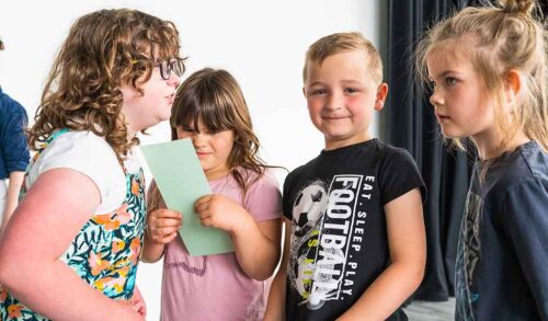 Four young children two on either end are looking at each other a girl in the middle is frowning at a piece of paper and a young boy in the middle is looking at the camera smiling