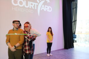 Four people are tightly squeezed into a large hula hoop, a session leader wearing a yellow t-shirt is looking at them smiling
