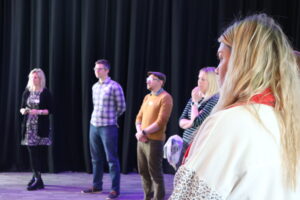 A group of people stand in a room with a grey floor, behind them is a black theatre curtain