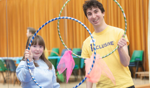 A young girl with downs syndrome stands next to a man wearing a yellow tshirt with inclusive written on it They are both holding large hula hoops