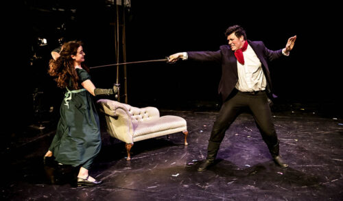 Stage Combat Workshop with Lydia  A girl with long dark hair wearing a green dress is in the middle of a sword fight with a man dressed in a suit and red neck scarf In the background there is a white chaise lounge