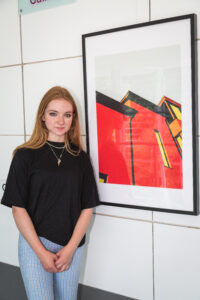 Artist Beth Gibson stands next to her artwork. It is a framed illustration of red buildings