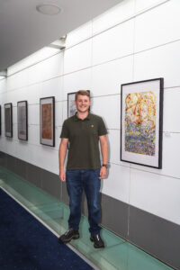 Artist Charles Gundy standing in front of his artwork, in large frames hanging on a wall