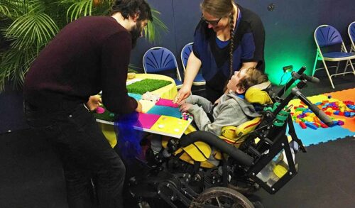 A young man in a wheelchair is taking part in craft activities facilitated by two workshop practitioners