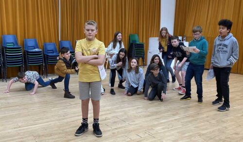 A young actor dressed in shorts and a yellow tshirt stands with his arms folded in a rehearsal room Behind him is a group of young performers posed in different positions