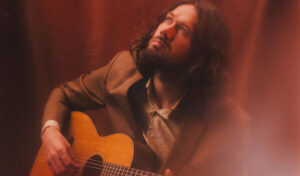 A close up of a man with long hair holding a guitar and looking upwards deep in thought