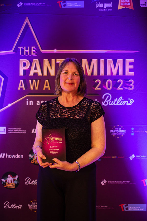 Director, Estelle van Warmelo, stood infront of a purple backdrop with UK Pantomime Awards written on it, holding the Best Director Award