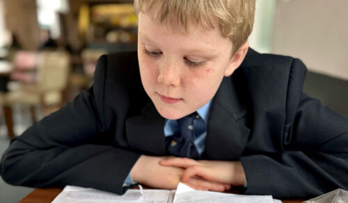 A young boy with blonde hair wearing a school uniform sits at a desk reading a book