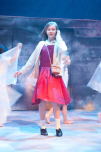 A young girl with shoulder length blonde hair stands on stage smiling. She is wearing a red dress and white cardigan and is tucking her hair behind her ear.