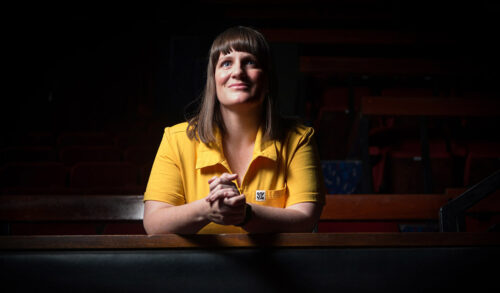 A woman with shoulder length brown hair sits in a pew with her hands clasped together She is wearing a bright yellow top