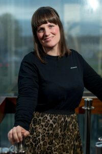 A woman with a long brown bob and heavy fringe stands smiling at the camera. She is wearing a black jumper and leopard print skirt