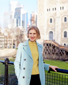 Royal historian Tracy Borman, a woman with a blonde bob wearing a yellow jumper under a pale blue coat leans against a fence in front of a castle