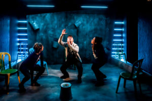 Tapped image: three performers on stage, two are crouched looking in on the middle performer who has his hands in the air and is looking up to the ceiling