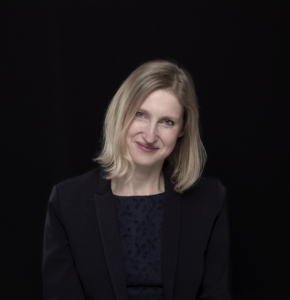 Tracy Borman, a woman with a short, blonde bob smiles at the camera. She is wearing a black top and blazer and is sitting against a black background
