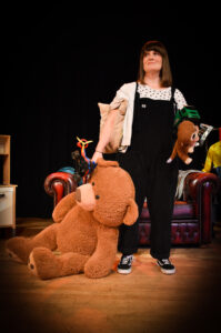 A woman wearing black dungarees over a white t-shirt stands holding a giant teddy bear in one hand, and a collection of children's toys in the other