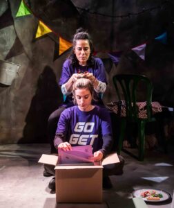 A blonde female actor sat on the floor in front of a cardboard box, reading some papers. She is wearing a purple t-shirt that reads 'go get'. Behind her is a a dark haired female performer who is playing with the other woman's hair