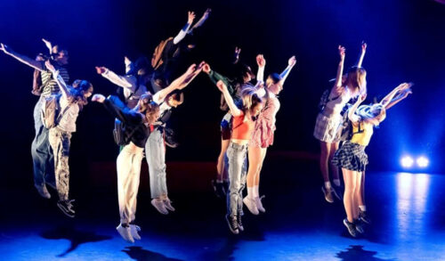 A group of dancers jumping int he air onstage They hold both arms in the air