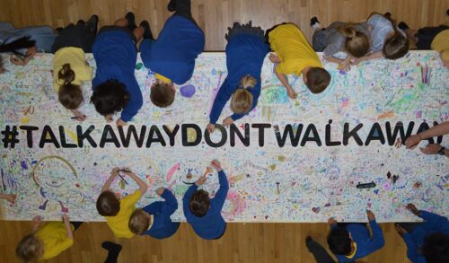 Children gathered around a table writing on a large piece of paper that reads TalkAwayDontWalkAway