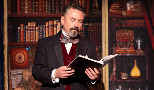 Image A man is stood in a library surrounded by books He is smartly dressed and reading a book