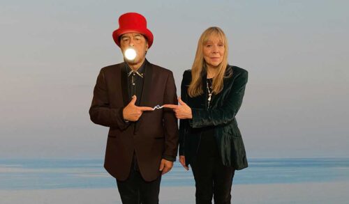 Image Two people stood in front of a blue background The man on the right has a lightbulb in his mouth and is wearing a red hat The lady on the left is dressed all in black They are about to touch their index fingers together there is a spark of electricity between them