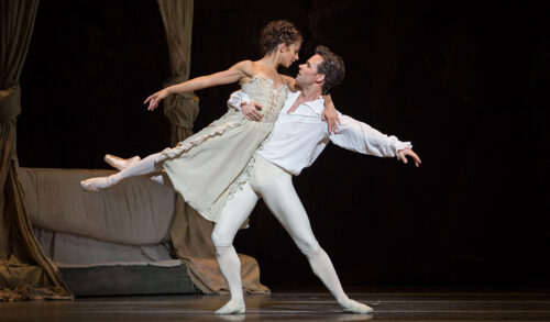 Two dancers on stage A man holds a woman with one arm lifting her in the air They look at each other