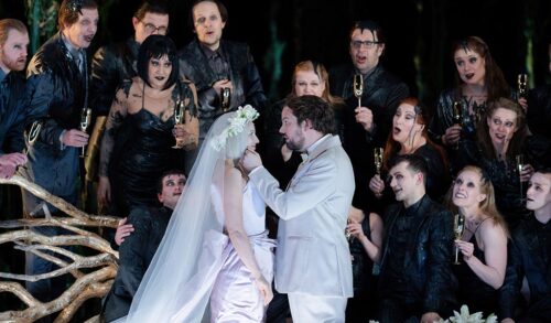 Two people kneel on stage getting married Surrounding them characters wearing all black look shocked