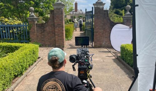 A man filming on a video camera through a gate of two actors walking away
