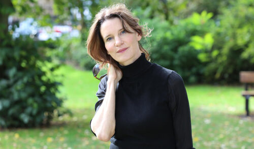 Image Susie Dent is stood in a park holding her neck and posing for the camera